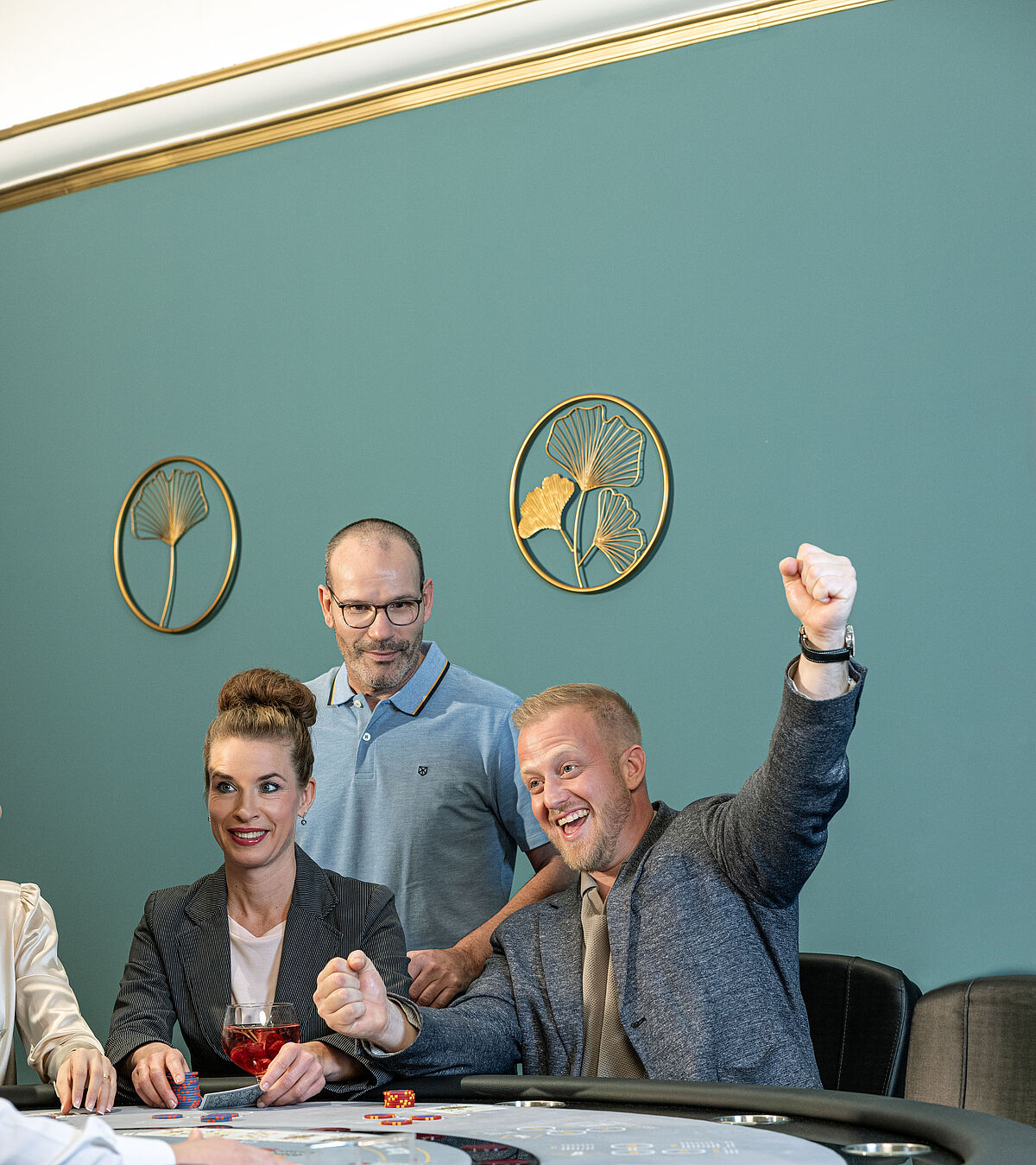 Eine Gruppe von Menschen versammelt sich um einen Casinotisch. Ein Mann rechts jubelt aufgeregt. Eine Frau hält lächelnd ein Getränk in der Hand. Ein Mann beobachtet das Spiel.