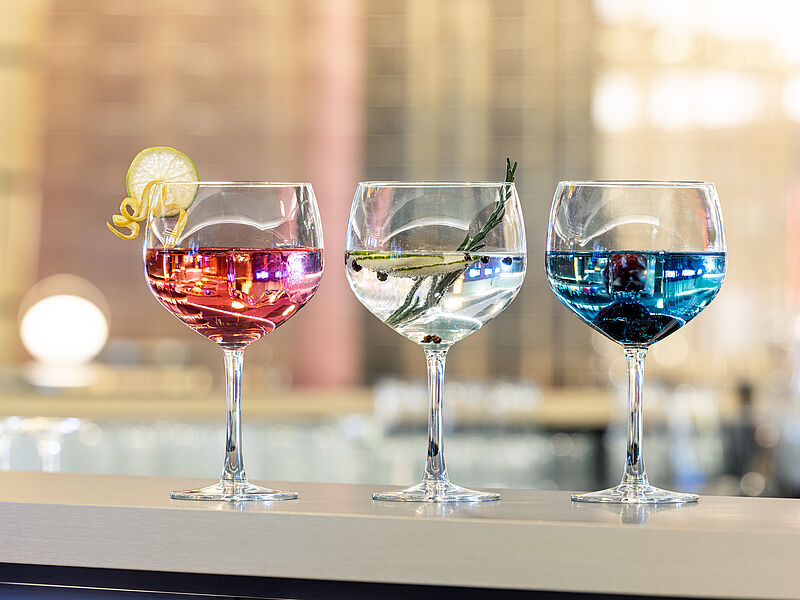 Drei elegante Gläser mit farbenfrohen Cocktails – rot mit einer Zitronenscheibe, klar mit Rosmarin und blau mit Beeren – stehen auf einer Bartheke und verströmen eine festliche Atmosphäre.