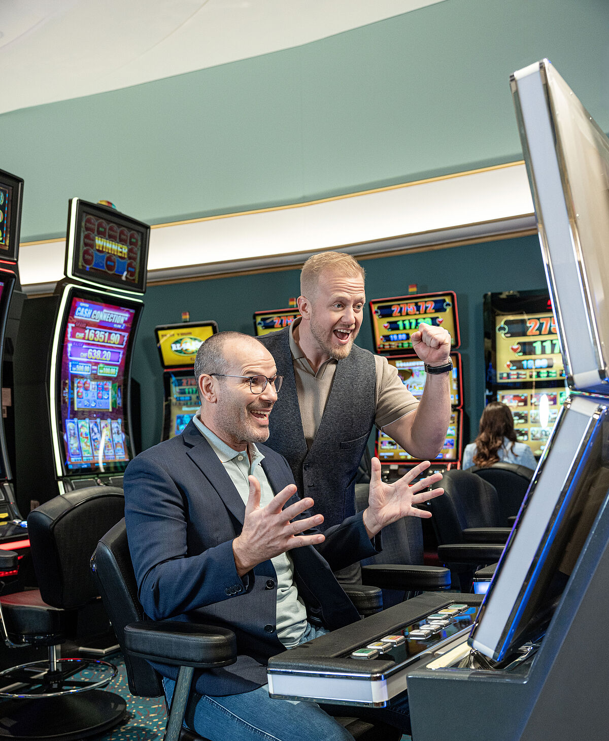 Zwei Männer in einem Casino spielen aufgeregt an einem Spielautomaten. Einer sitzt da und gestikuliert enthusiastisch, während der andere jubelnd dasteht und die Faust ballt.