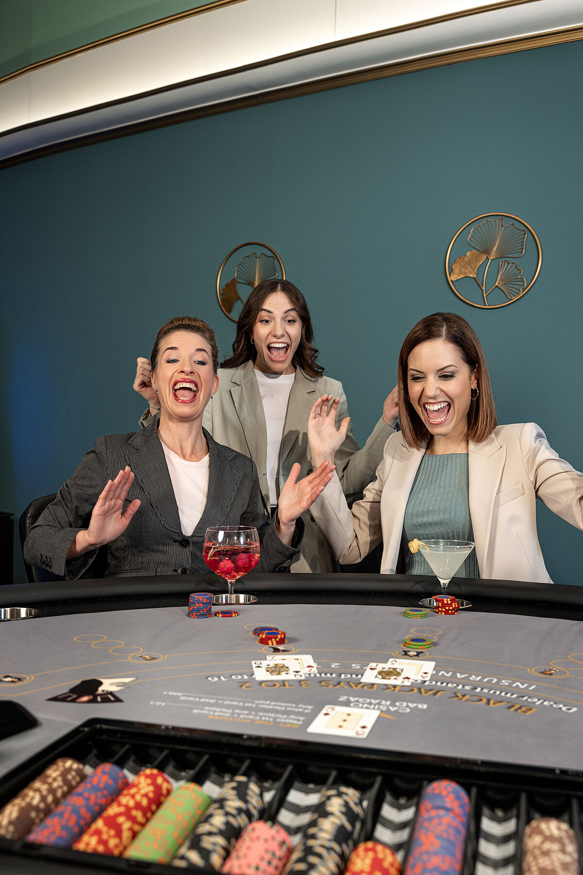 Drei Frauen an einem Casinotisch feiern ihren Gewinn. Sie lächeln und heben aufgeregt die Hände. Chips und Karten liegen auf dem Tisch.