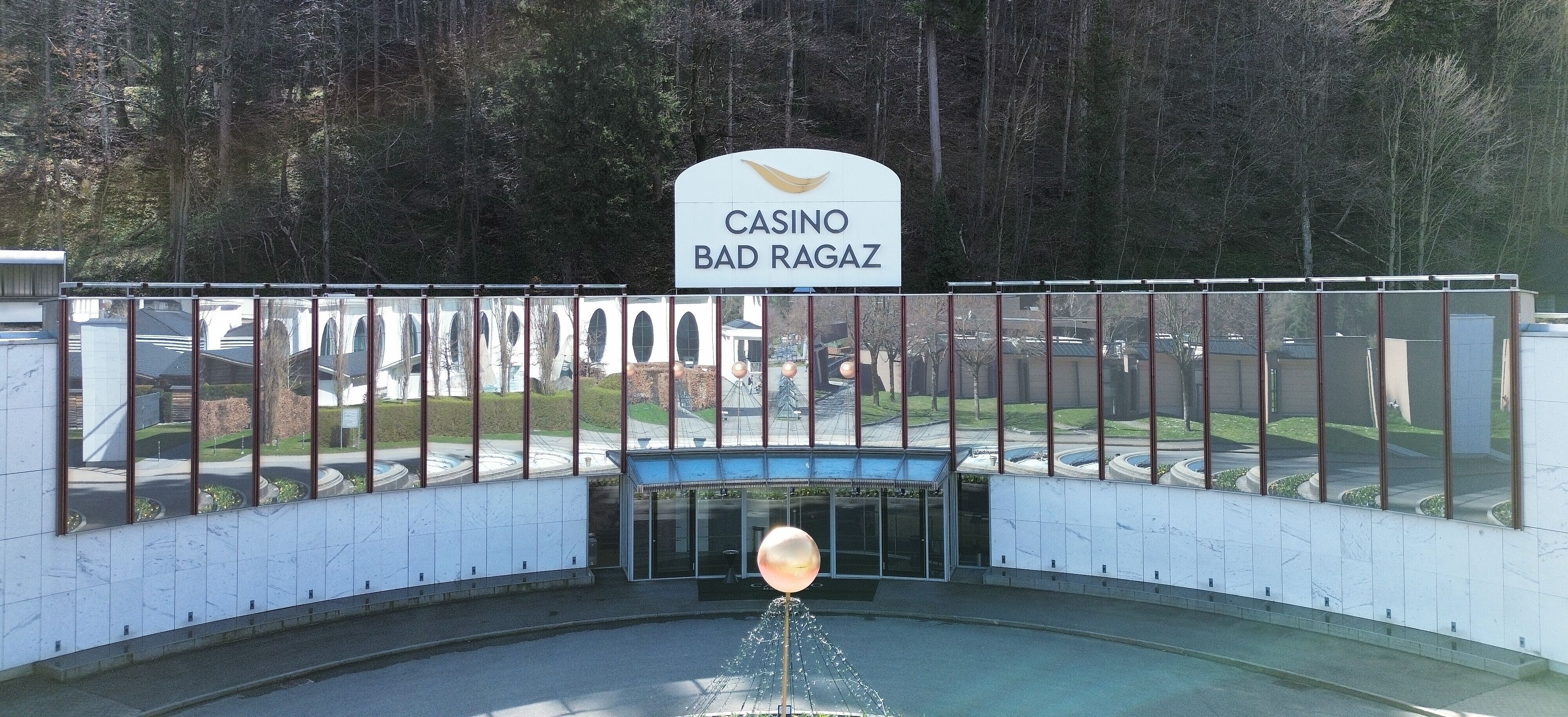  Aussenansicht des Casino Bad Ragaz mit grossen Spiegelfenstern, in denen sich Bäume spiegeln. Der moderne Eingang besticht durch einen leuchtenden Kugelbrunnen. Ruhige, einladende Atmosphäre.