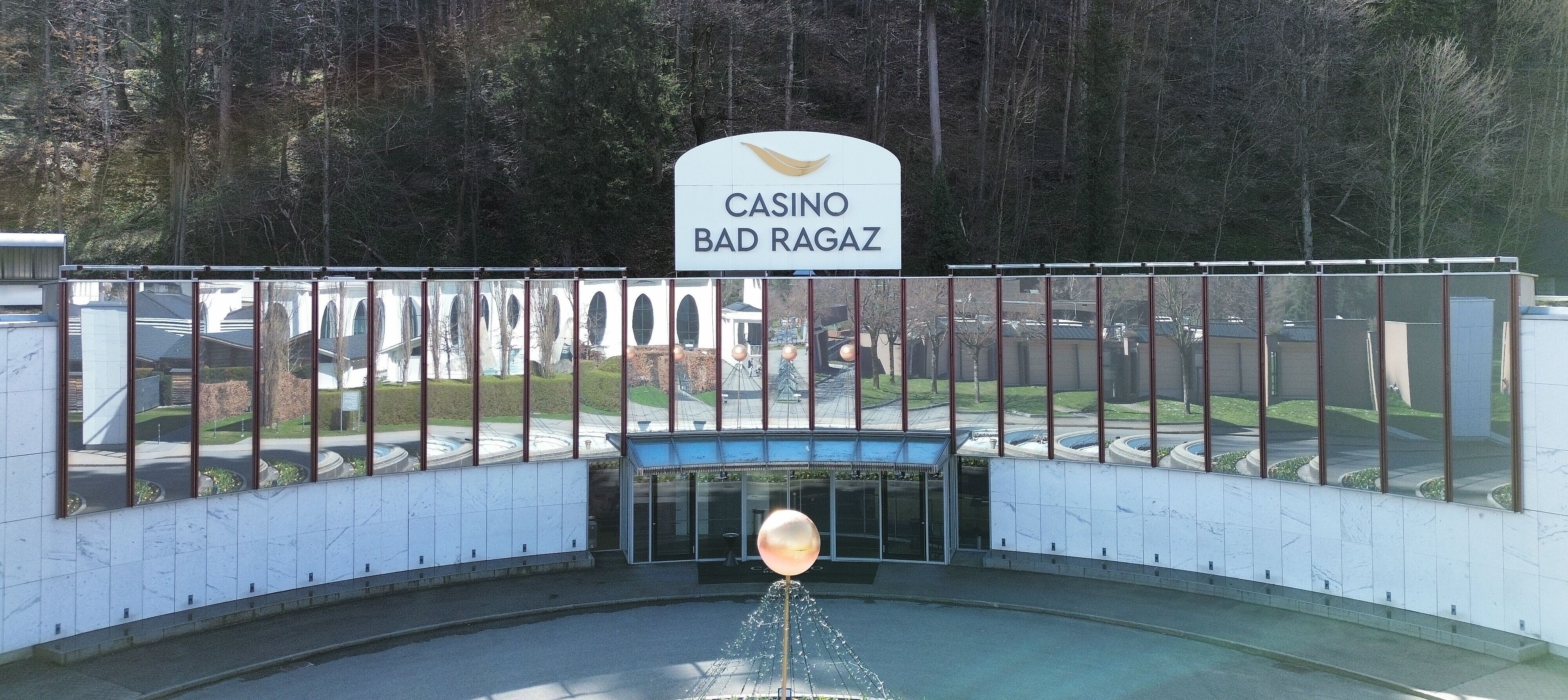  Aussenansicht des Casino Bad Ragaz mit grossen Spiegelfenstern, in denen sich Bäume spiegeln. Der moderne Eingang besticht durch einen leuchtenden Kugelbrunnen. Ruhige, einladende Atmosphäre.