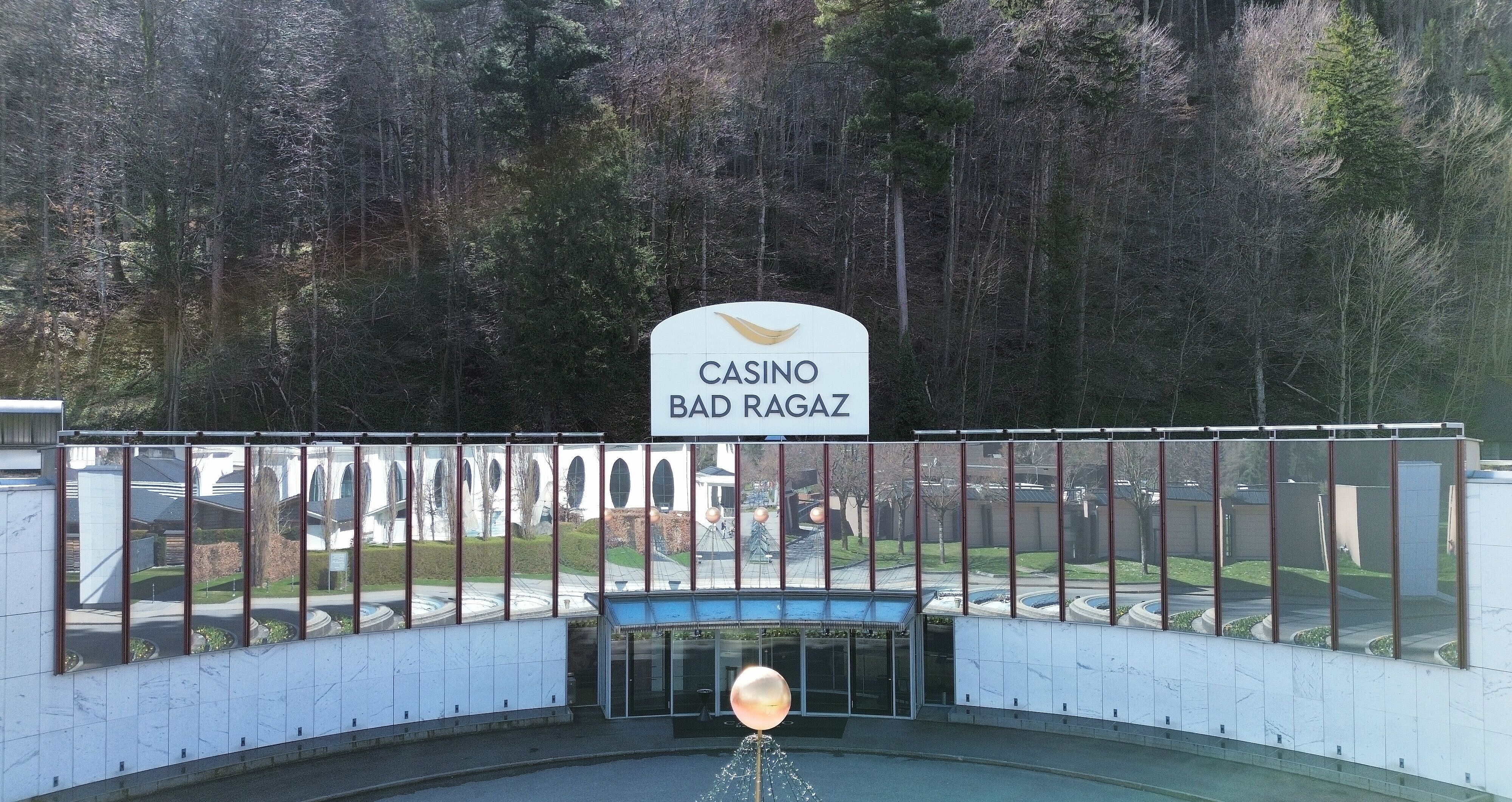  Aussenansicht des Casino Bad Ragaz mit grossen Spiegelfenstern, in denen sich Bäume spiegeln. Der moderne Eingang besticht durch einen leuchtenden Kugelbrunnen. Ruhige, einladende Atmosphäre.