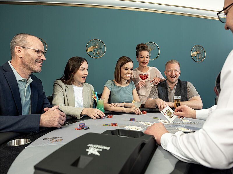  Menschen versammelten sich aufgeregt um einen Pokertisch. Ein Dealer verteilt Karten. Chips liegen verstreut auf dem Tisch und deuten auf ein spannendes Spiel hin.