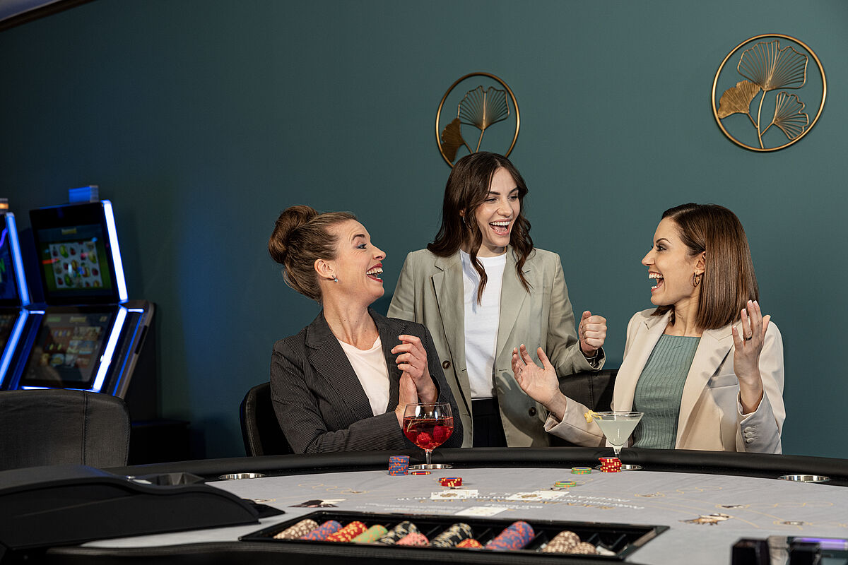 Drei Frauen feiern in einem Casino an einem Spieltisch mit Jetons. Sie drücken Aufregung und Freude aus und deuten auf einen Gewinn hin. In der Nähe leuchten Spielautomaten.