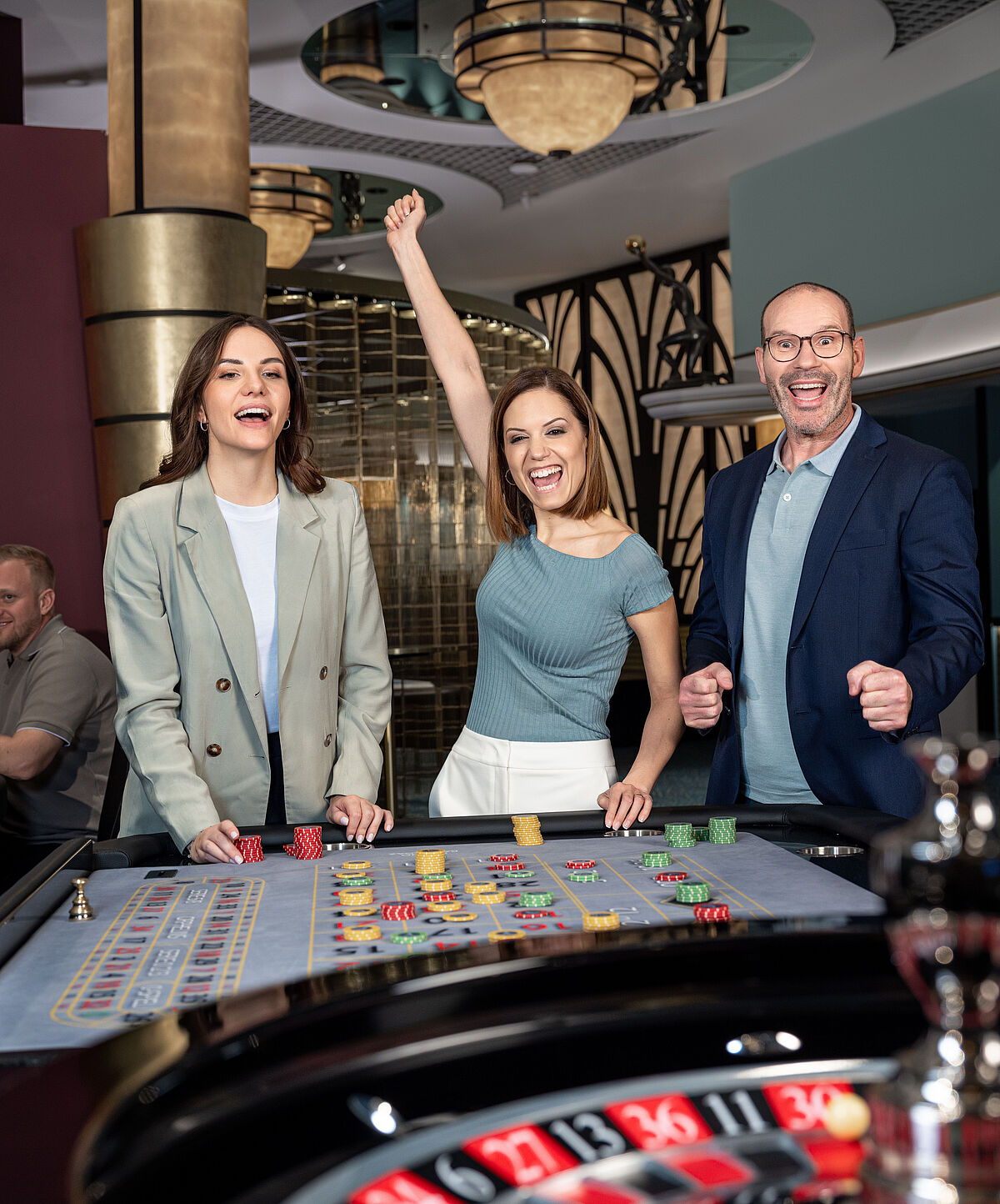  Drei Personen jubeln an einem Casino-Roulettetisch. Eine Frau streckt aufgeregt den Arm hoch und sorgt so für eine ausgelassene, feierliche Atmosphäre.
