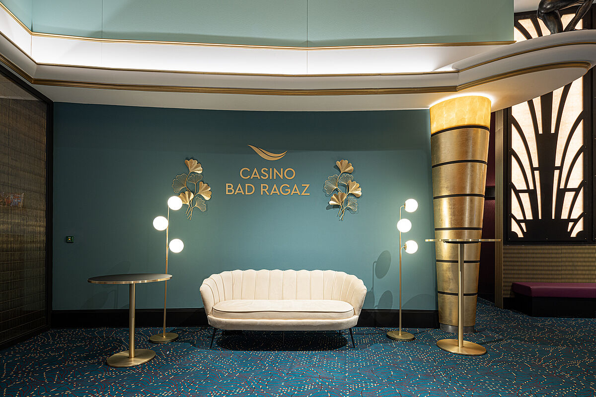  Elegante Casino-Lobby mit blaugrünen Wänden, goldenen Akzenten, einem weissen Sofa, modernen Lampen und runden Tischen. Anspruchsvolle, einladende Atmosphäre.