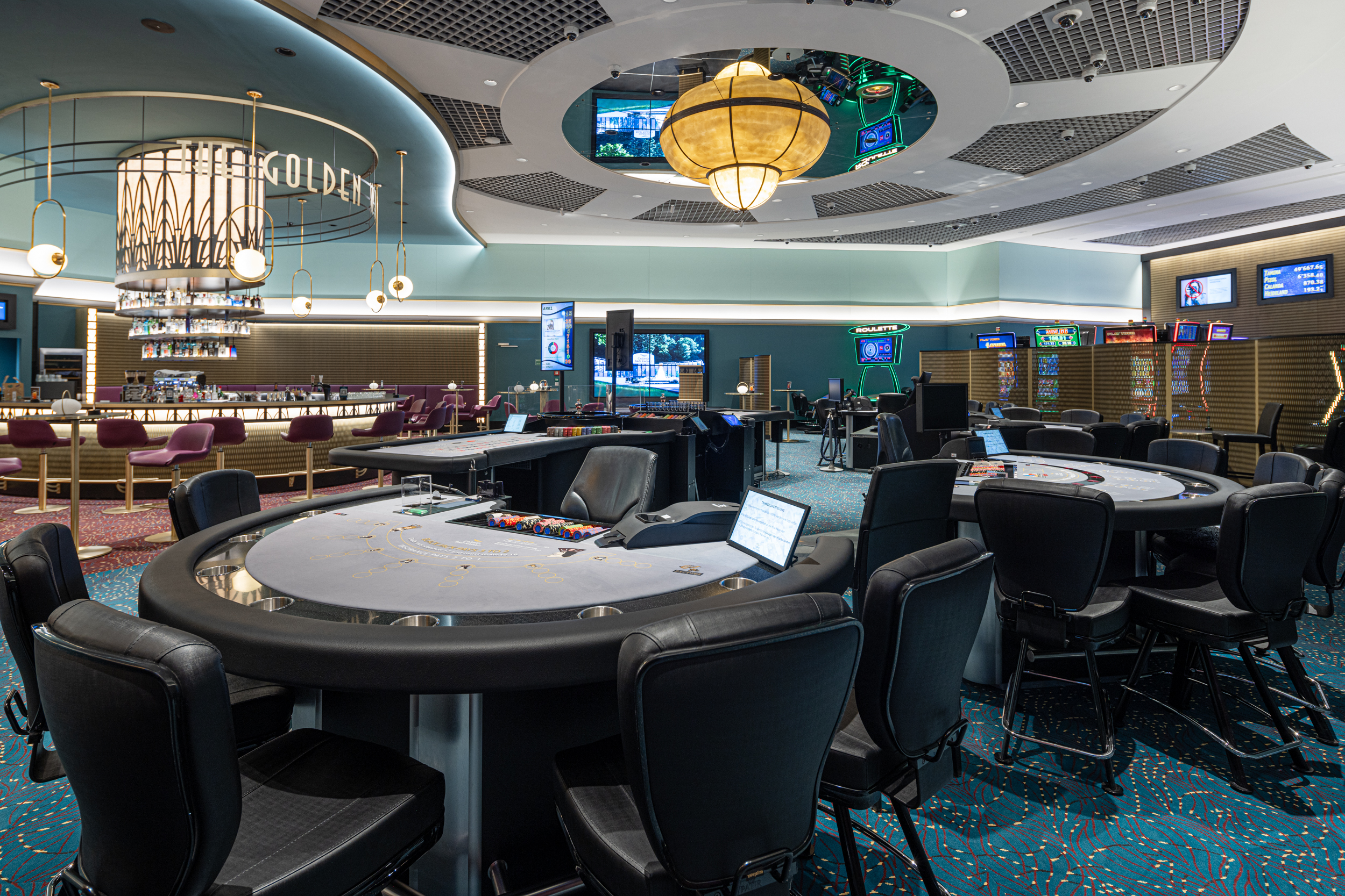 Modernes Casino-Interieur mit eleganten schwarzen Spieltischen und Stühlen, stimmungsvoller Beleuchtung, dekorativer Decke und Bildschirmen mit Spielinformationen; lebhafte Atmosphäre.