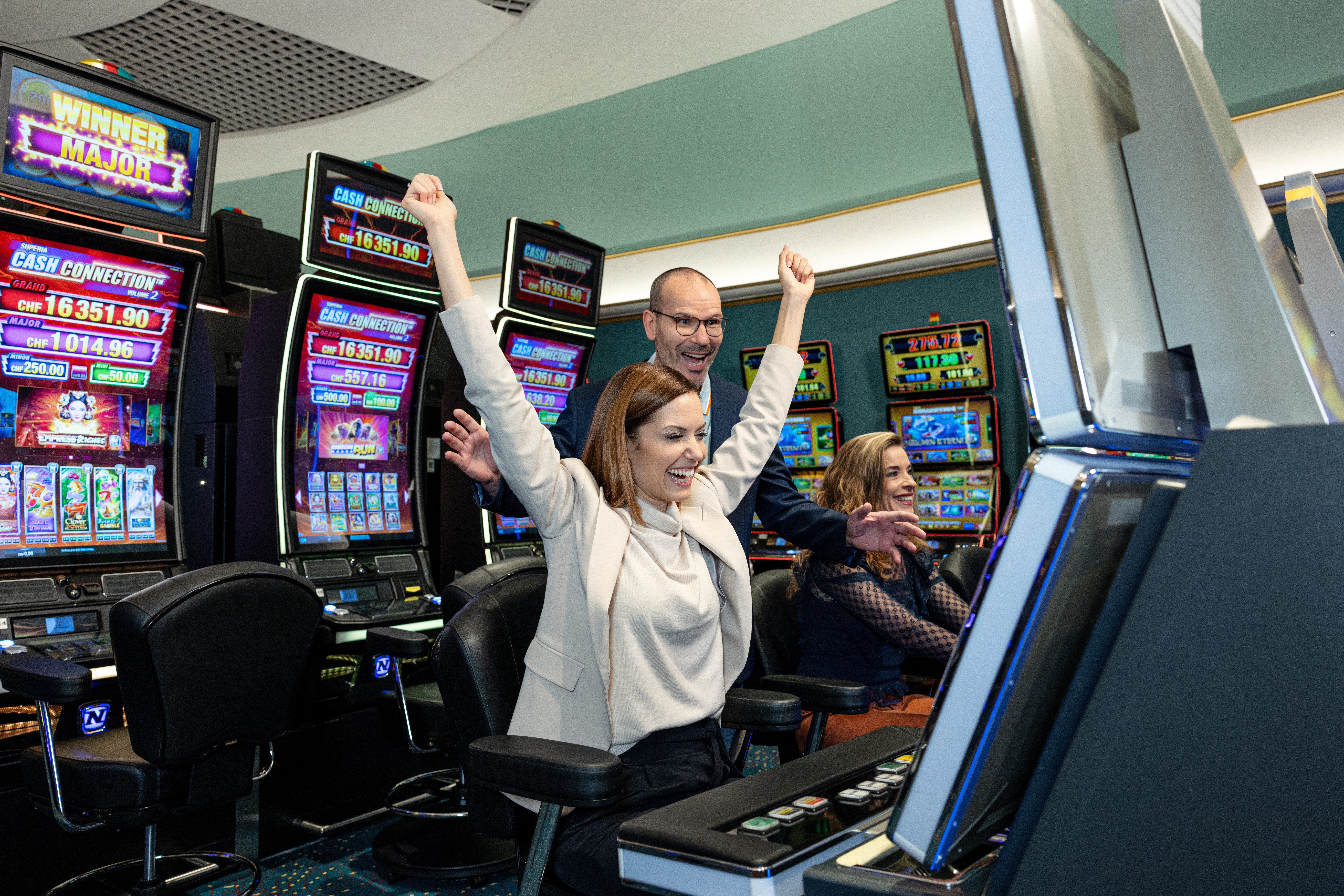 Eine Frau reckt freudig die Arme in die Höhe, während sie vor einem Spielautomaten in einem Casino sitzt. Im Hintergrund sind bunte Bildschirme und andere Gäste zu sehen.
