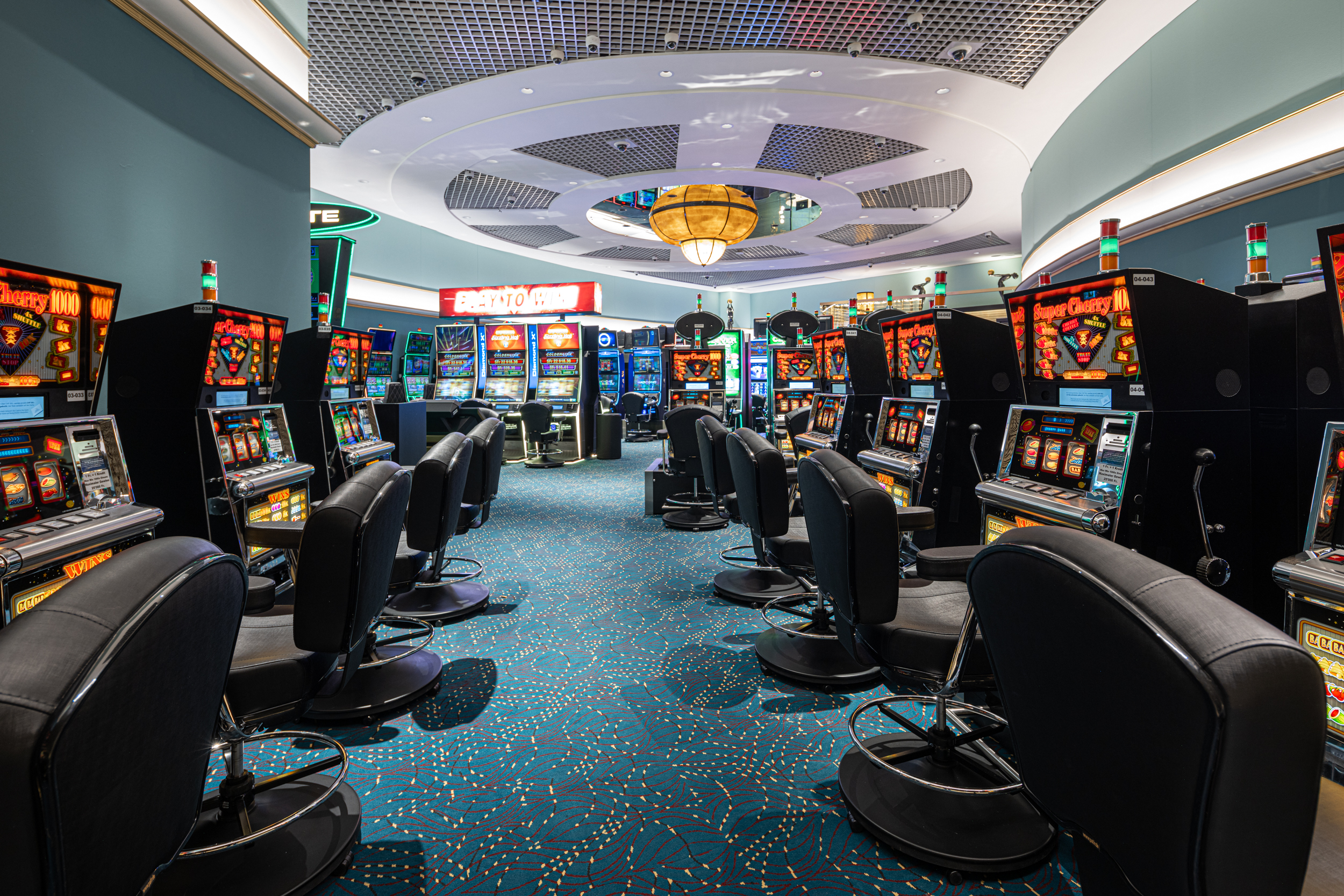  Ein lebendiges Casino-Interieur mit Reihen von Spielautomaten mit leuchtenden Bildschirmen und bequemen schwarzen Stühlen auf einem gemusterten blauen Teppich unter kreisförmiger Beleuchtung.