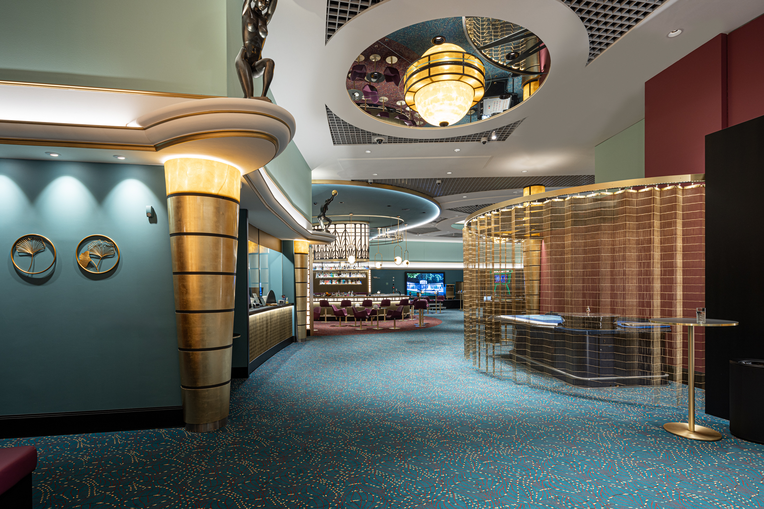Luxuriöse Lobby mit türkisfarbenem Teppich, goldenen Akzenten und modernen Kronleuchtern. Skulpturen und runde Spiegel an der Decke schaffen eine elegante Atmosphäre.