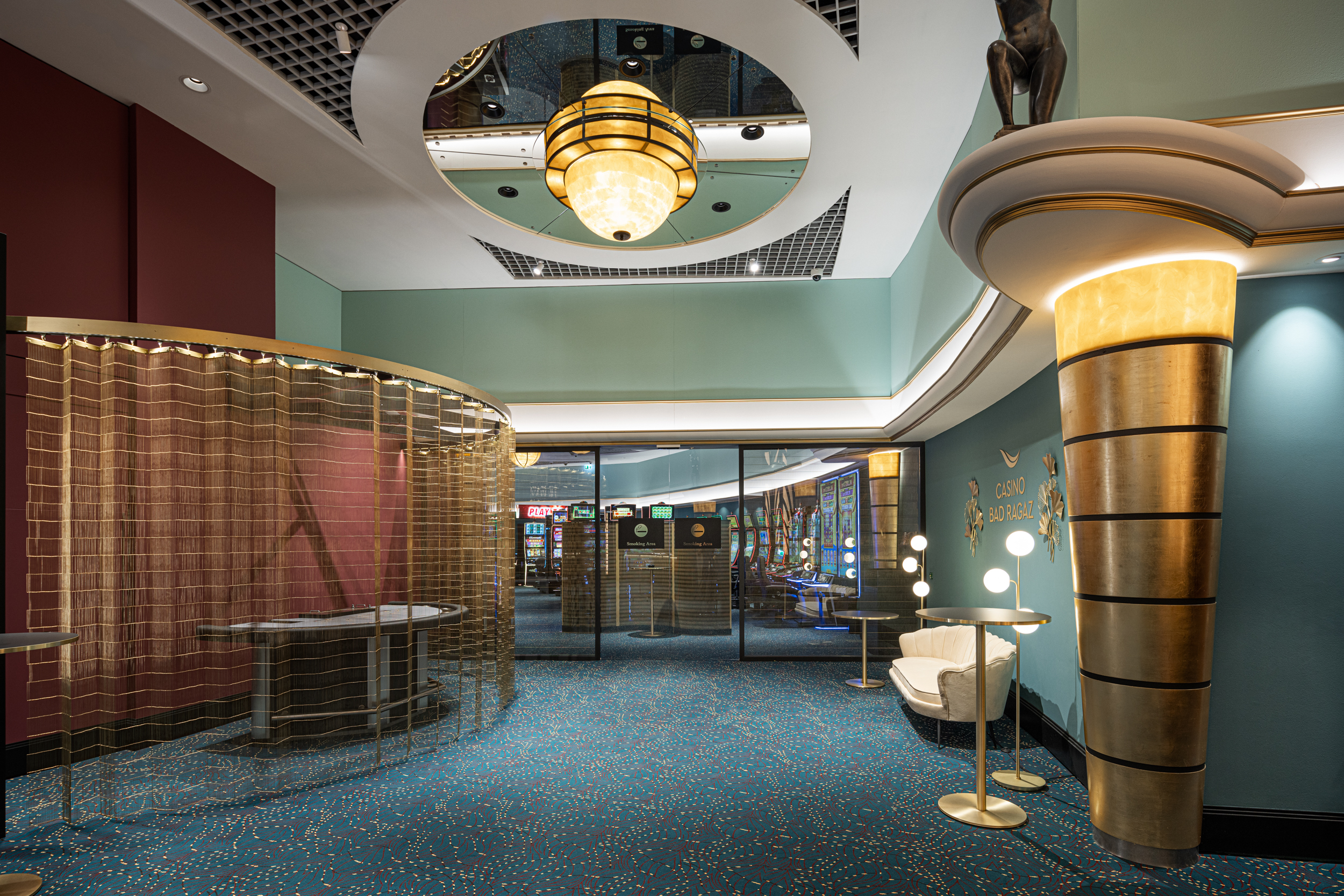 Lobby im Art-déco-Stil mit lebendigem Dekor. Verfügt über einen blauen Teppich mit bunten Punkten, goldenen Akzenten, einzigartiger Beleuchtung und einem Deckenspiegel, in dem sich ein kunstvoller Kronleuchter spiegelt.