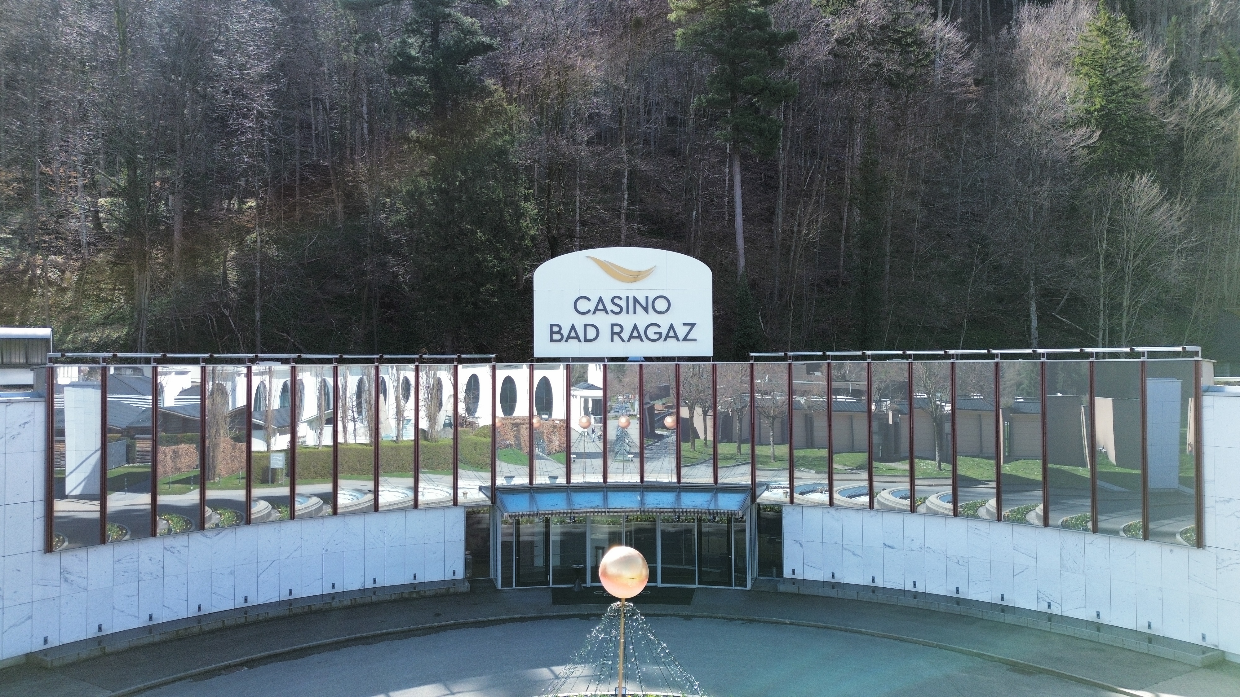  Aussenansicht des Casino Bad Ragaz mit grossen Spiegelfenstern, in denen sich Bäume spiegeln. Der moderne Eingang besticht durch einen leuchtenden Kugelbrunnen. Ruhige, einladende Atmosphäre.